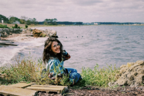 Ellery sits near the ocean, facing the camera in a blue jacket