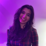 Cameron is smiling and surrounded by purple lighting. She is wearing a red striped dress.