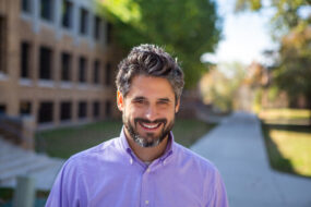 Man with salt and pepper hair in a blue-purple shirt behind a brick building