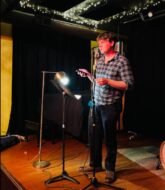 A man in a plaid shirt and dark jeans stands with a music stand and light in front of him reading